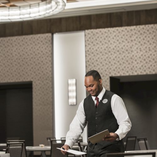 A person in a formal vest is setting up tables in a spacious, modern conference room with patterned walls and a unique ceiling light.