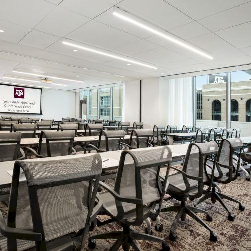 A modern classroom or conference room with rows of chairs and tables, large windows, and a screen displaying "Texas A&M Hotel and Conference Center."