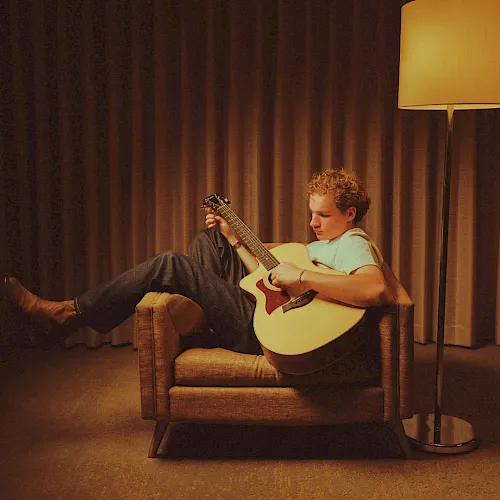 A boy sits on a couch playing an acoustic guitar, with warm lamp light and curtains behind him.