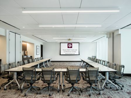 A modern conference room features a U-shaped table setup, mesh office chairs, and a presentation screen with Texas A&M branding.