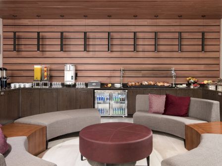 A cozy lounge with curved seating and cushions, featuring beverage dispensers, snacks, and a fridge. Warm wood paneling accents the space.