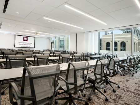 A conference room with rows of tables and chairs facing a screen displaying "Texas A&M Hotel and Conference Center" by large windows.