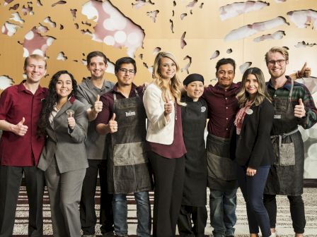 A group of nine people standing in a row, smiling and giving thumbs up, wearing a mix of business and casual attire.