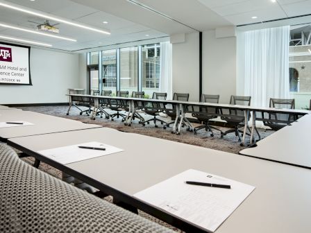 A modern conference room with tables, chairs, notepads, and a screen reading "Texas A&M Hotel and Conference Center."