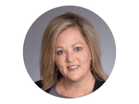 The image shows a person with light hair and a neutral expression, wearing a dark top and earrings, against a gray background.