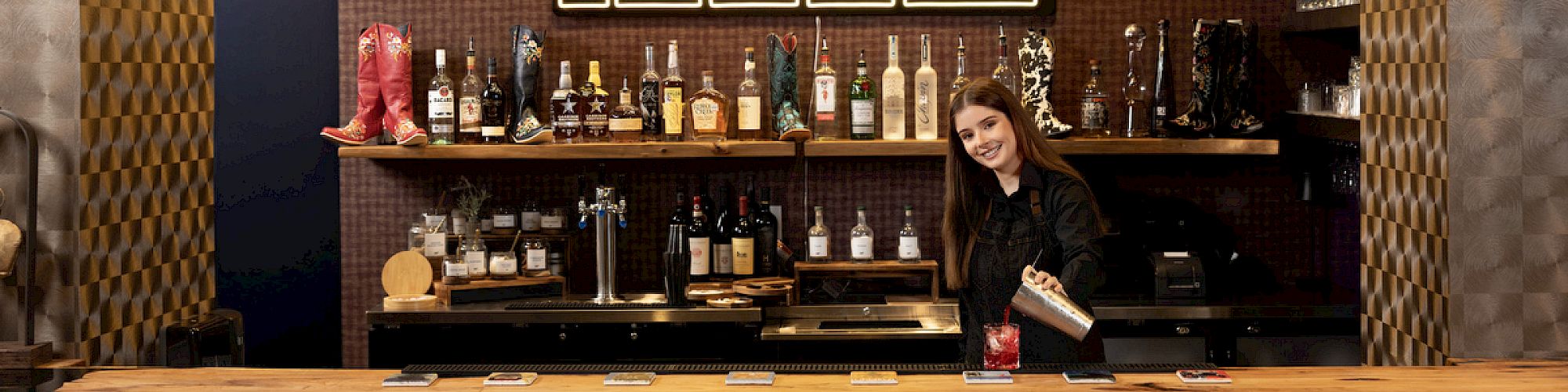 A bar with a wooden counter, various bottles on shelves, and a person behind the counter beneath a "HOWDY" sign.