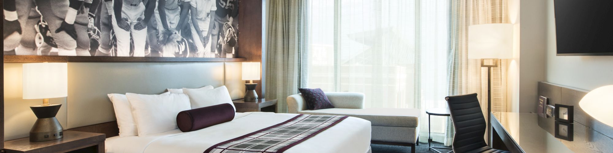 The image shows a modern hotel room with a large bed, a sports-themed picture, a desk, a TV, and a window with sheer curtains.