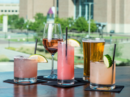 An assortment of colorful drinks including cocktails, wine, and beer, served on napkins with various garnishes, set against an urban backdrop.