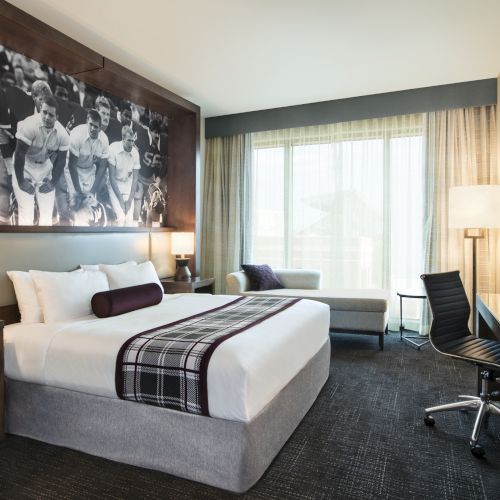 A modern hotel room with a large bed, desk, chair, and wall art of a football scene, featuring natural light from a large window.