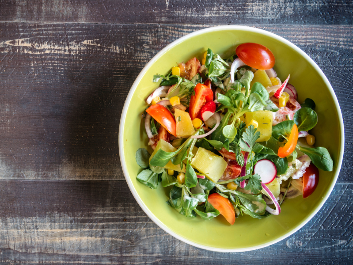 A colorful salad in a green bowl with tomatoes, corn, leafy greens, radishes, and bell peppers on a rustic wooden table.