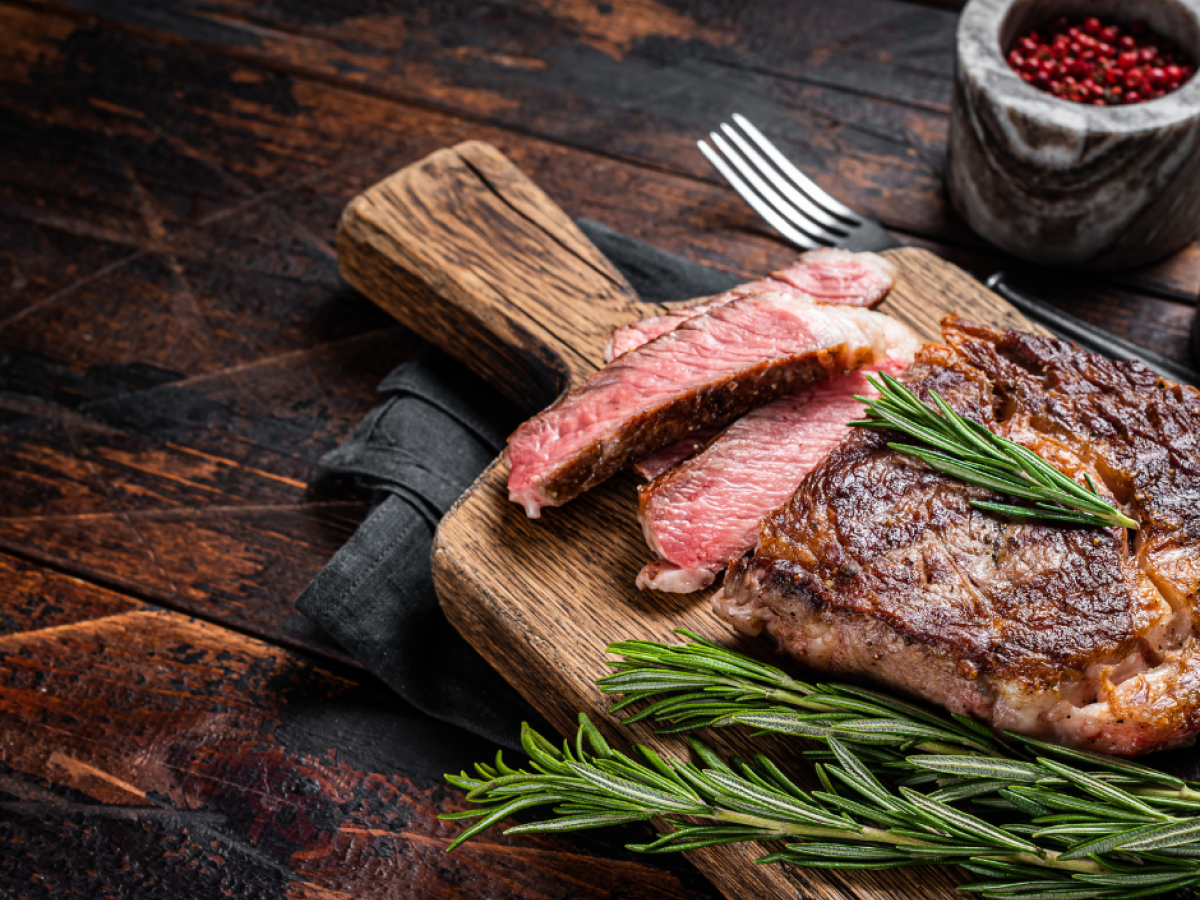 A juicy steak, sliced medium-rare, is on a wooden board with sprigs of rosemary.