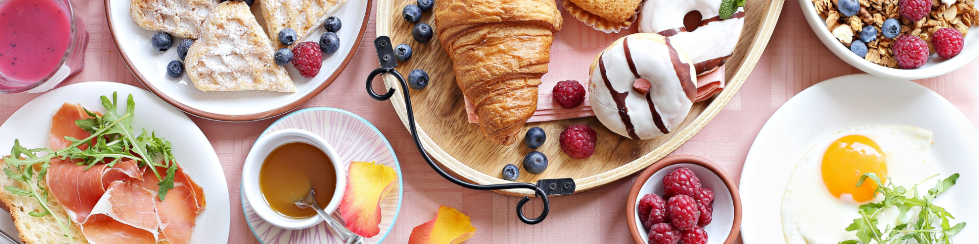 A breakfast spread with waffles, croissant, muffin, yogurt parfait, fried egg, toast with ham, berries, juice, and coffee on a pink table.