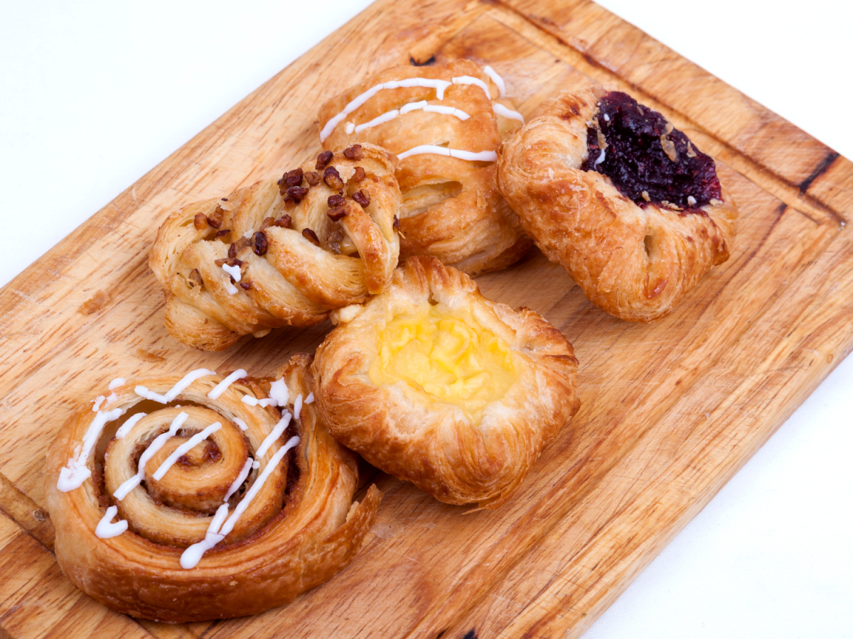 The image shows various pastries, including a cinnamon roll and fruit-filled danishes, arranged on a wooden cutting board with a white background.