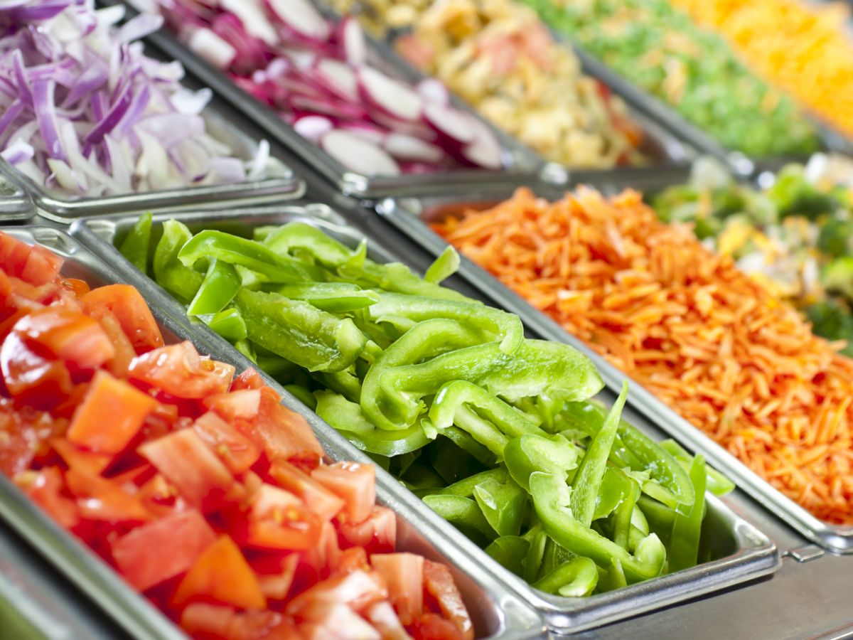 A colorful salad bar with chopped red bell peppers, green bell peppers, shredded carrots, purple onions, and other fresh ingredients.