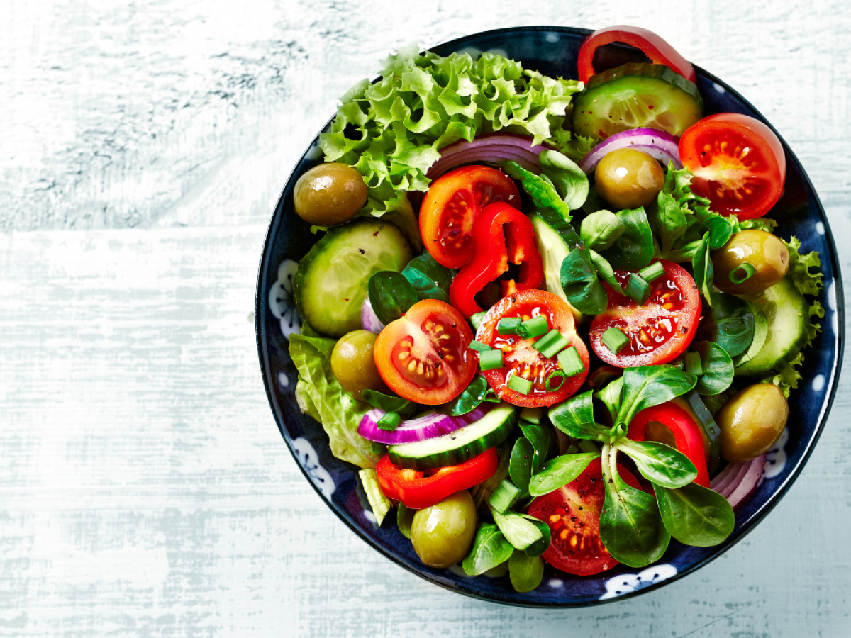 A vibrant salad with lettuce, tomatoes, cucumbers, olives, red onion, red bell pepper, and greens on a dark plate.