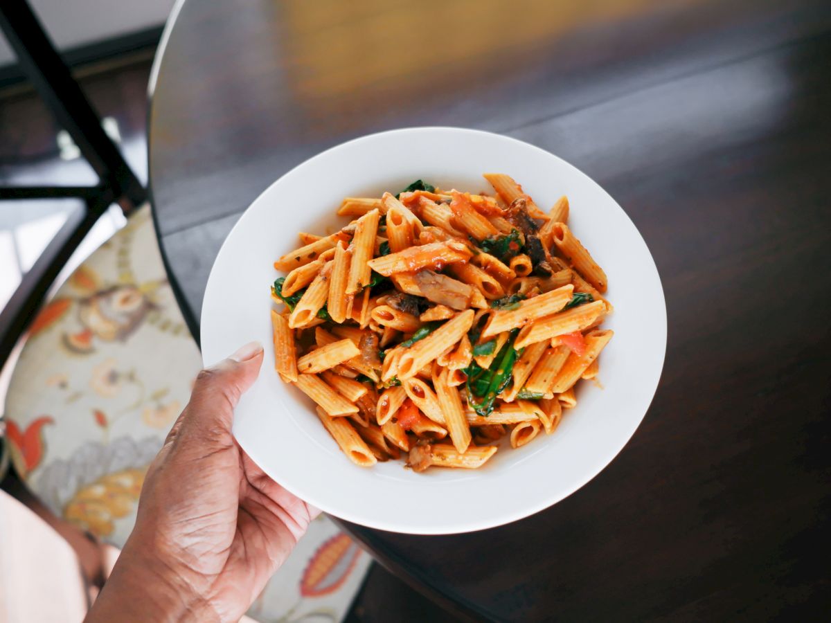 A bowl of penne pasta tossed with tomato sauce, herbs, and greens, held over a dark table by a hand. Bon app&eacute;tit.