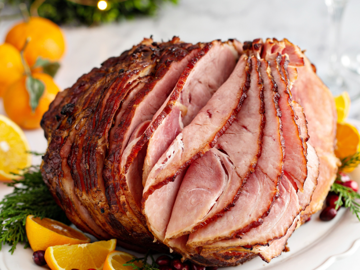 A glazed, sliced ham garnished with orange slices and cranberries, set on a festive table decorated with greenery and small oranges.