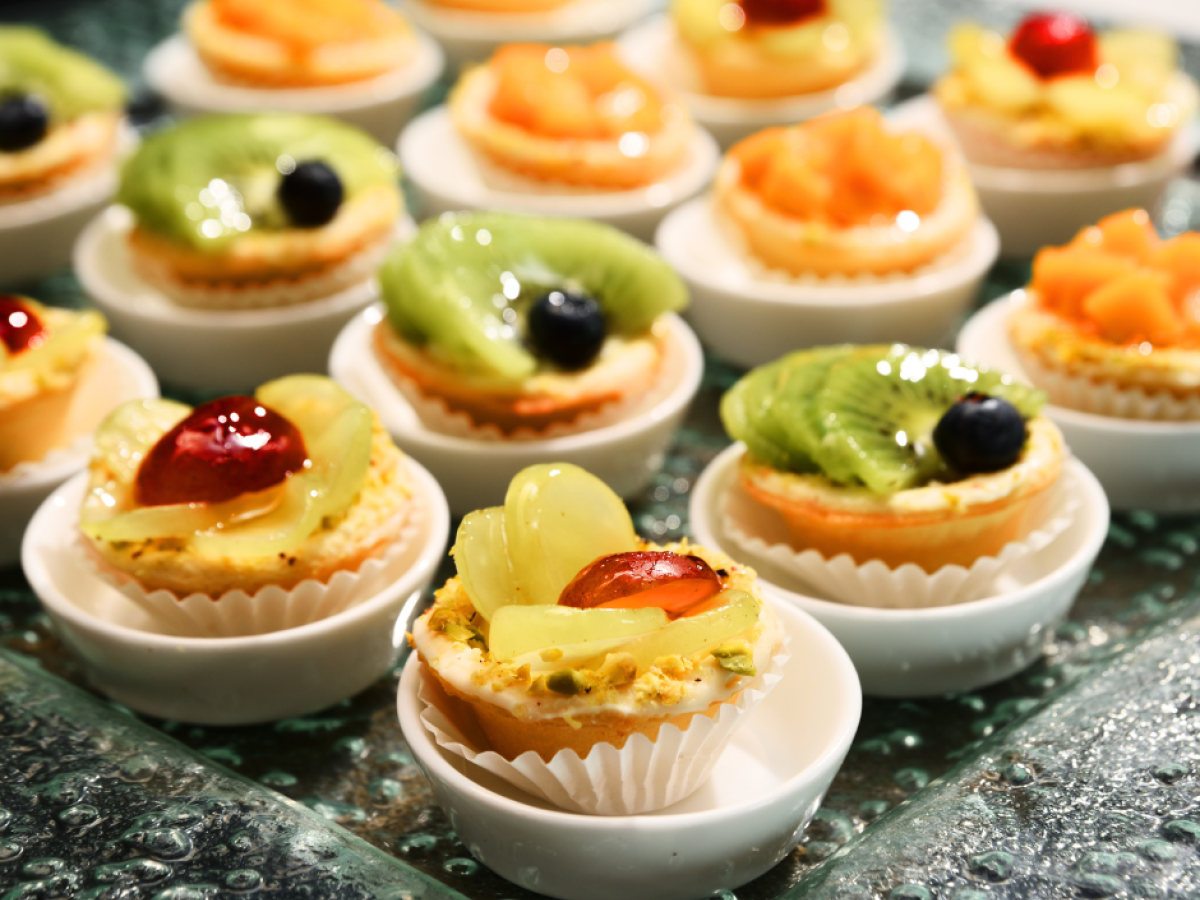 Mini fruit tarts are displayed, featuring kiwis, grapes, and berries on a decorative tray in small white cups.