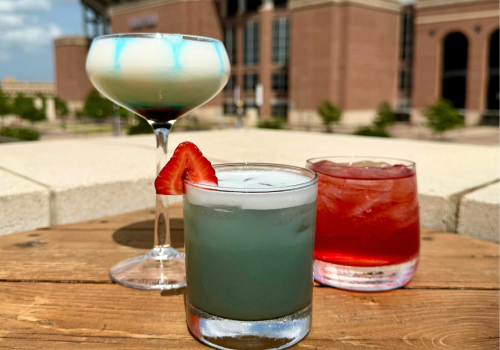 Three colorful cocktails on a wooden table with a stadium in the background.