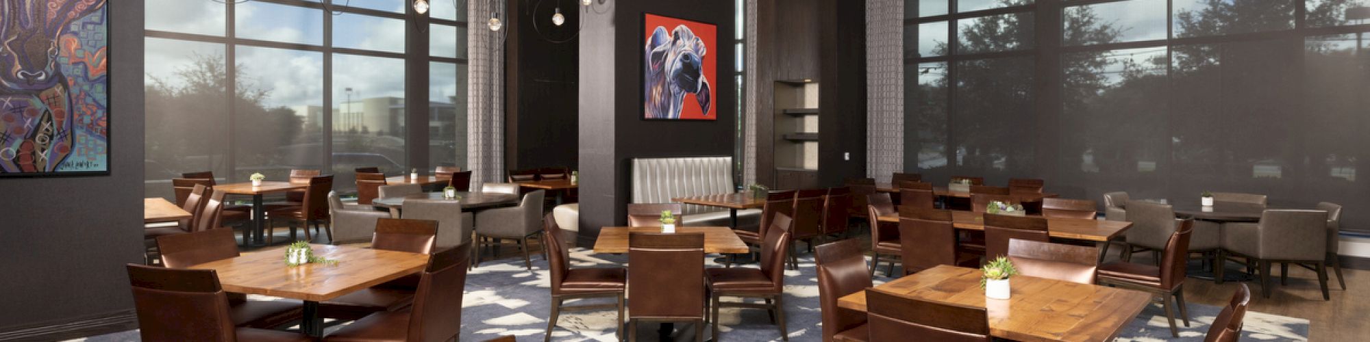 The image shows a modern restaurant interior with wooden tables, brown chairs, large windows, and colorful artwork on the walls.