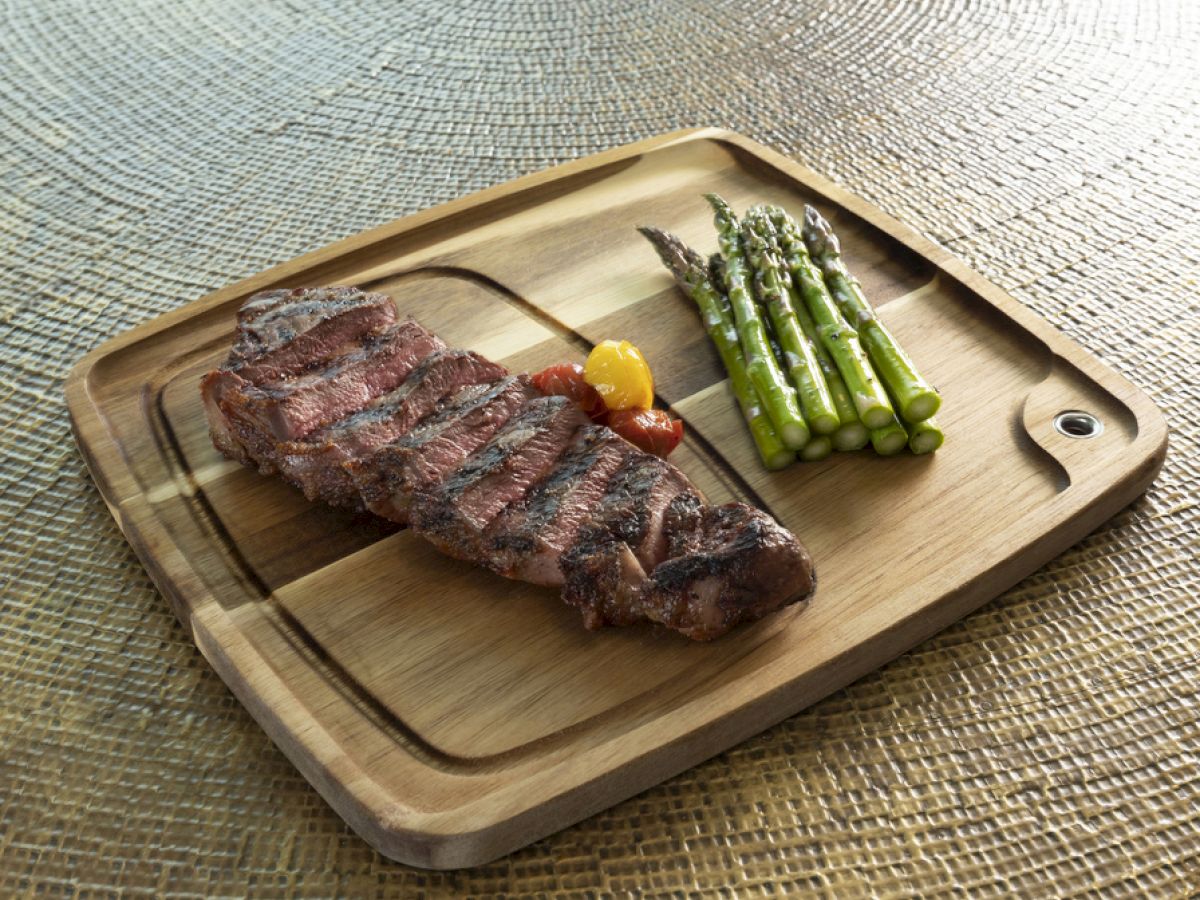 A wooden tray featuring a grilled steak with asparagus and a small pepper, set on a textured surface.
