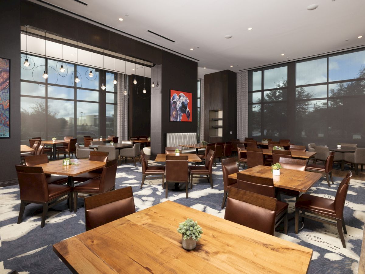 A contemporary dining area with wooden tables, chairs, abstract art on walls, modern lighting, and large windows illuminating the space.