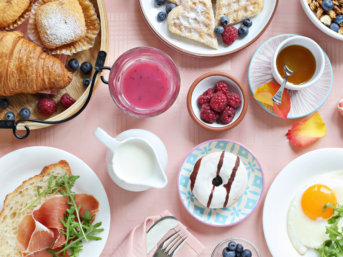 A breakfast spread with croissants, waffles, fruit, yogurt, juice, toast with ham, and a sunny-side-up egg on a pink cloth.