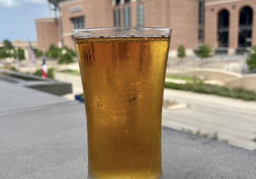 A glass of amber liquid, possibly beer, sits on a surface with a large building and clear sky in the background.
