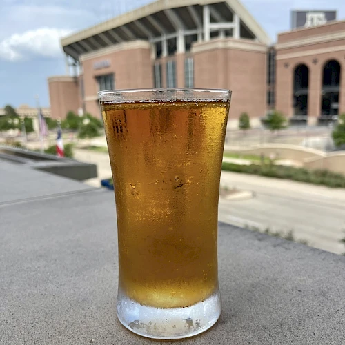 A glass of amber liquid, possibly beer, sits on a surface with a large building and clear sky in the background.