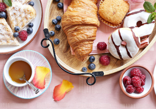 A variety of breakfast foods: croissants, donuts, waffles, egg, berries, yogurt, granola, ham, bread, and a drink on a pink tablecloth.