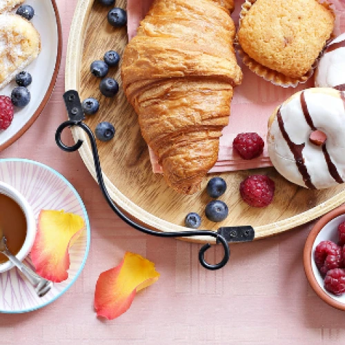 A variety of breakfast foods: croissants, donuts, waffles, egg, berries, yogurt, granola, ham, bread, and a drink on a pink tablecloth.