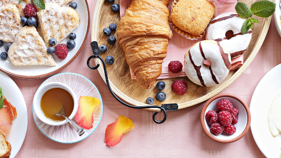 A variety of breakfast foods: croissants, donuts, waffles, egg, berries, yogurt, granola, ham, bread, and a drink on a pink tablecloth.