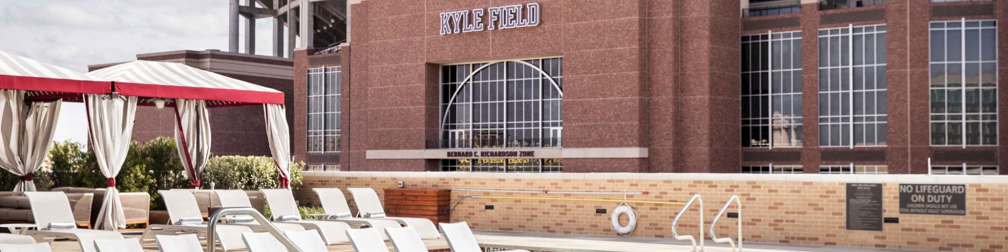 A swimming pool with lounge chairs is in the foreground, and a large stadium named "Kyle Field" is in the background.