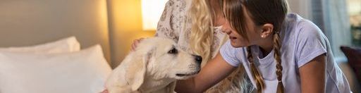 A blonde woman and a girl sit on a bed with a fluffy puppy, sharing a joyful moment indoors, lights warm and cozy, smiles all around.
