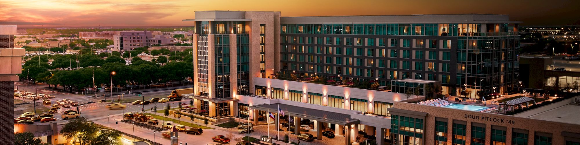 A modern building complex at sunset, featuring a hotel with expansive windows, surrounded by landscaped paths and streetlights.