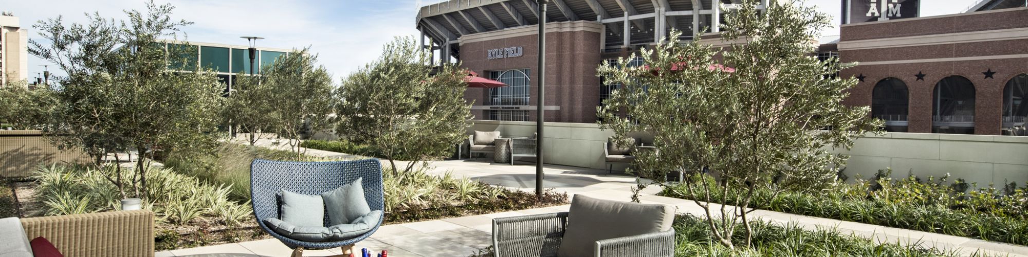 An outdoor seating area with sofas and chairs is set near a stadium, surrounded by greenery under a blue sky.