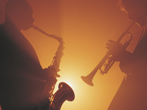 Silhouettes of two musicians playing a saxophone and a trumpet against an orange-lit background.
