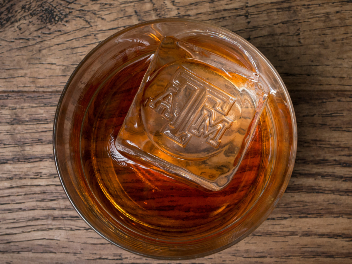 A glass of amber-colored liquid with an ice cube imprinted with "ATM" on a wooden surface.