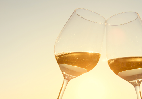 Two stemmed wine glasses filled with amber-gold liquid, catching warm golden light against a pale background.