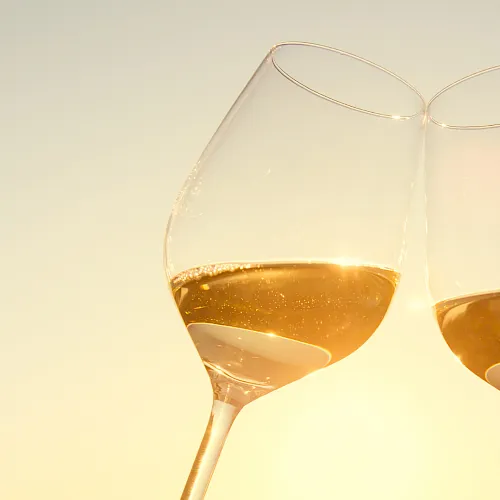 Two stemmed wine glasses filled with amber-gold liquid, catching warm golden light against a pale background.