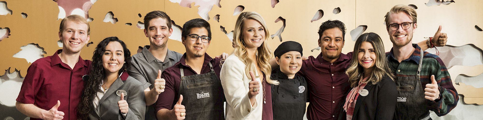 A group of nine people, some in work aprons and others in business casual attire, smile and give thumbs-ups in front of a decorated wall.