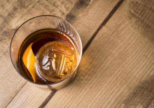 A glass of whiskey with ice and an orange slice on a wooden table, viewed from above.