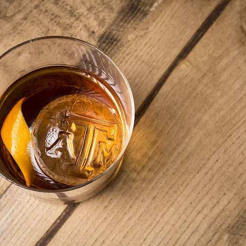 A glass of whiskey with ice and an orange slice on a wooden table, viewed from above.