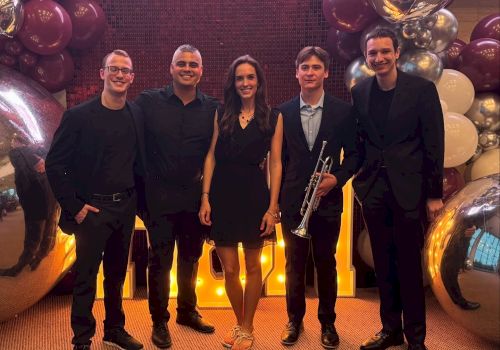 A group of five people posing together; one holds a trumpet. They're surrounded by large reflective balloons and lights.