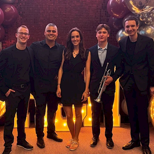 A group of five people posing together; one holds a trumpet. They're surrounded by large reflective balloons and lights.