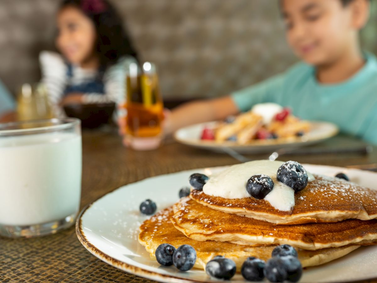 A plate of pancakes topped with blueberries and cream, a glass of milk, and two kids blurred in the background enjoying breakfast.