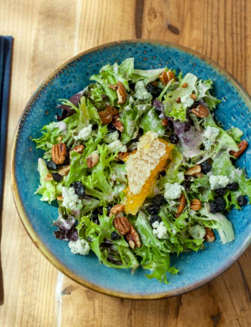 A fresh salad with mixed greens, pecans, and cheese on a blue plate; utensils and a drink are set beside on a wooden table.
