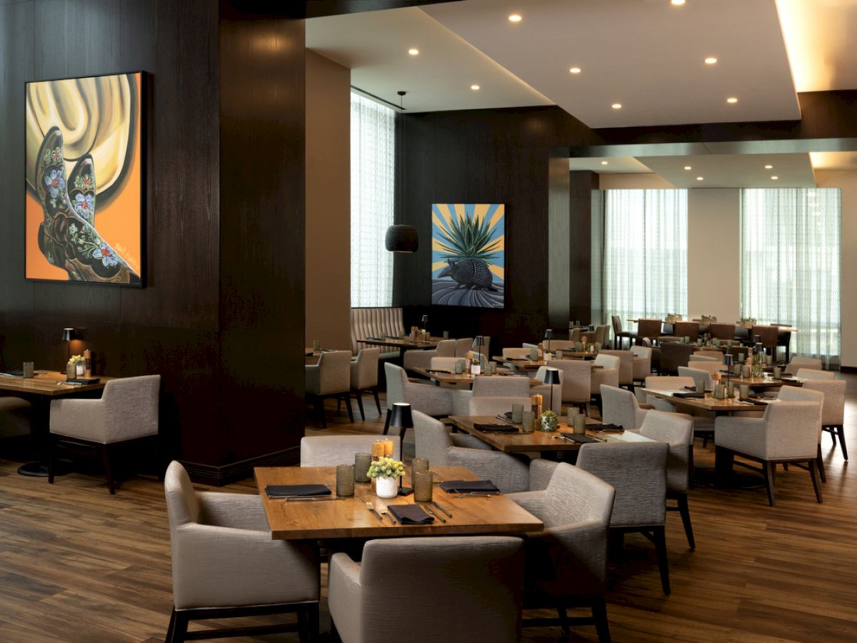 A modern restaurant with dark walls, wooden floors, and art on the walls; gray upholstered chairs surround wooden tables set with candles.