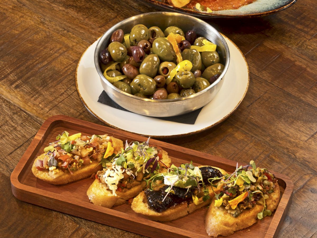 The image shows an arrangement of dishes including roasted vegetables, a bowl of olives, and a platter of assorted bruschetta on a wooden table.