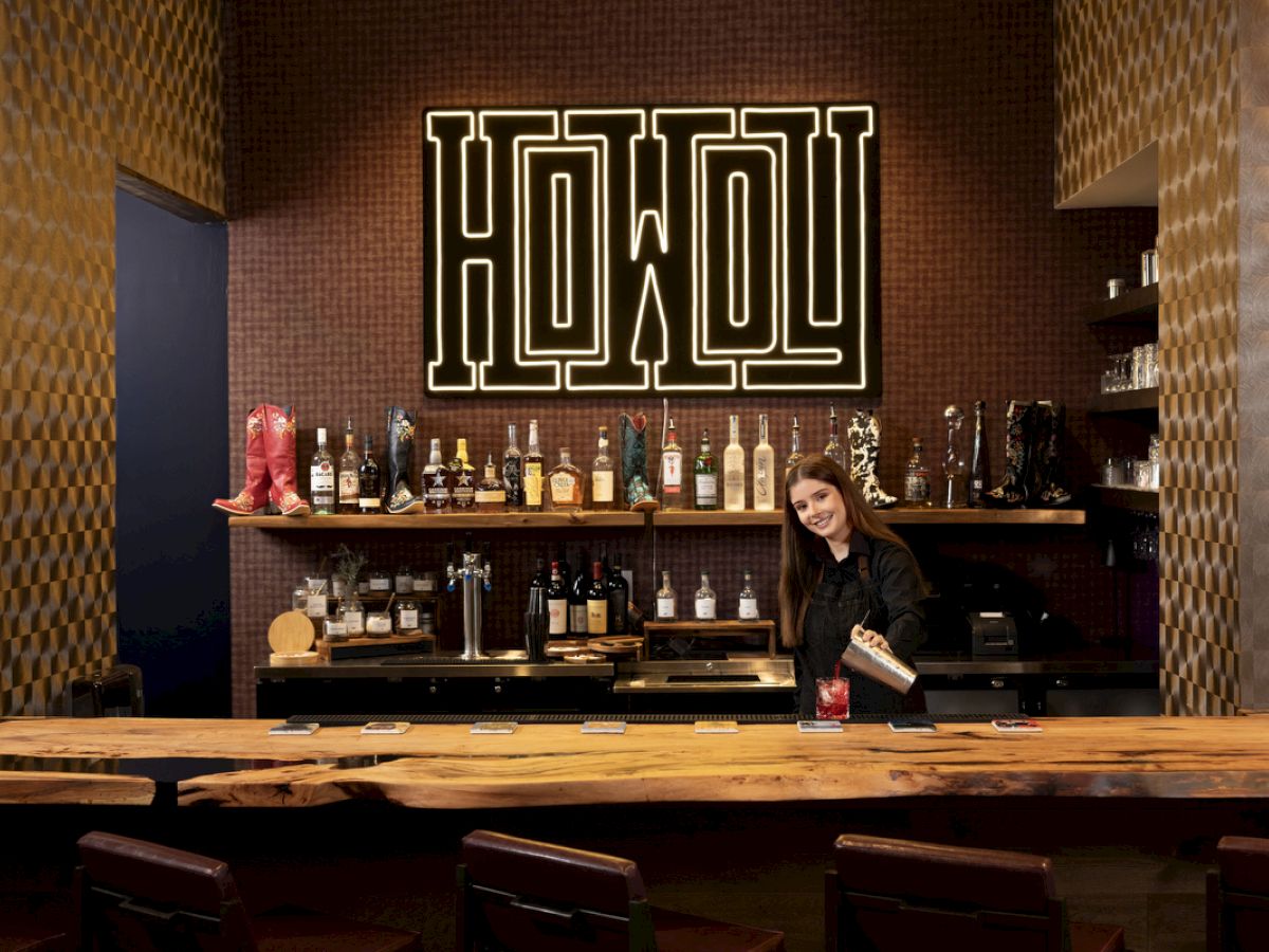 A bar scene with a person preparing a drink, surrounded by various bottles, beneath a large sign reading "HOWDY".